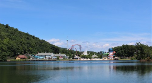 A scenic view of an amusement park by a lake, featuring rides and lush green hills under a clear blue sky.
