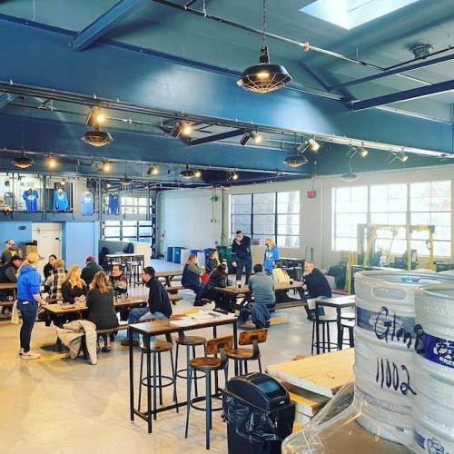 A spacious indoor venue with people seated at tables, enjoying food and drinks, under industrial-style lighting.