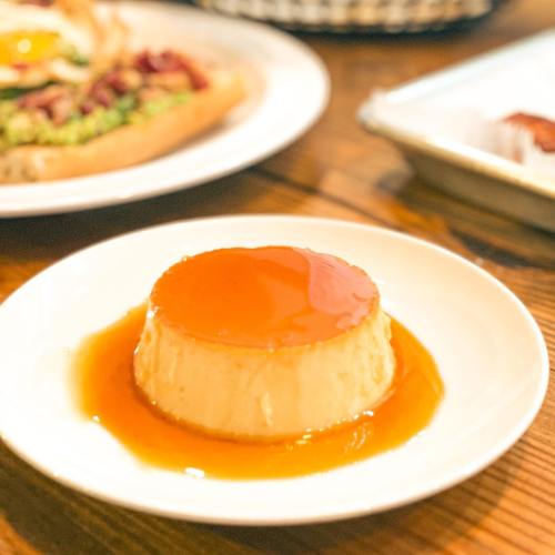 A creamy flan topped with caramel sauce, served on a white plate with a blurred background of food.