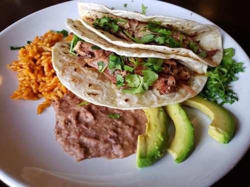 Three tacos filled with meat, topped with cilantro, served with rice, refried beans, and avocado slices on a white plate.