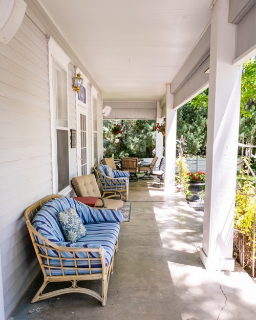 A cozy porch with wicker chairs and cushions, surrounded by greenery and colorful flowers. Sunlight filters through.