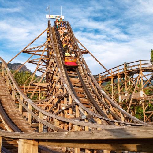 A roller coaster climbs a steep wooden track with riders enjoying the thrill against a blue sky.