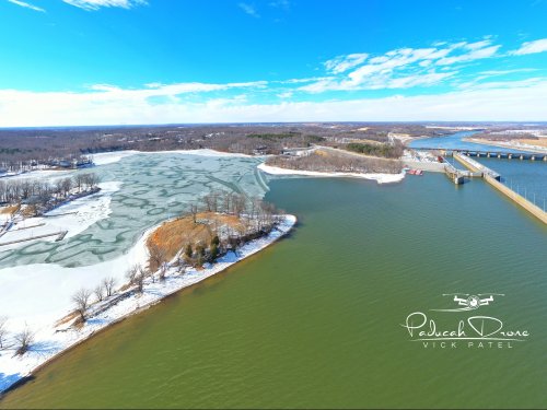 Aerial view of a frozen lake with snow-covered banks, surrounded by trees and a river under a clear blue sky.