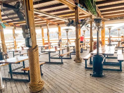 A spacious outdoor dining area with wooden tables, fans, and heaters, overlooking a waterfront view.