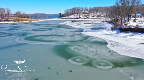 Aerial view of a frozen lake with circular ice patterns and trees along the shore under a clear blue sky.