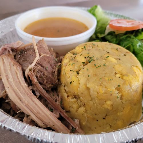 A plate featuring shredded meat, a side of yellow mashed plantains, salad, and a small cup of broth.