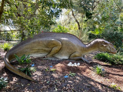 A life-sized dinosaur sculpture lying near a nest of eggs, surrounded by lush greenery in a garden setting.