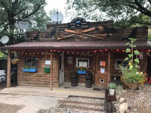 A rustic wooden building with a sign, satellite dishes, and flower pots, serving as a store for camping and groceries.
