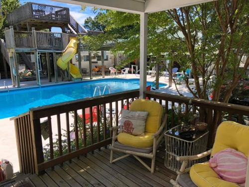 A sunny backyard with a pool, water slide, and seating area featuring yellow chairs and a view of greenery.