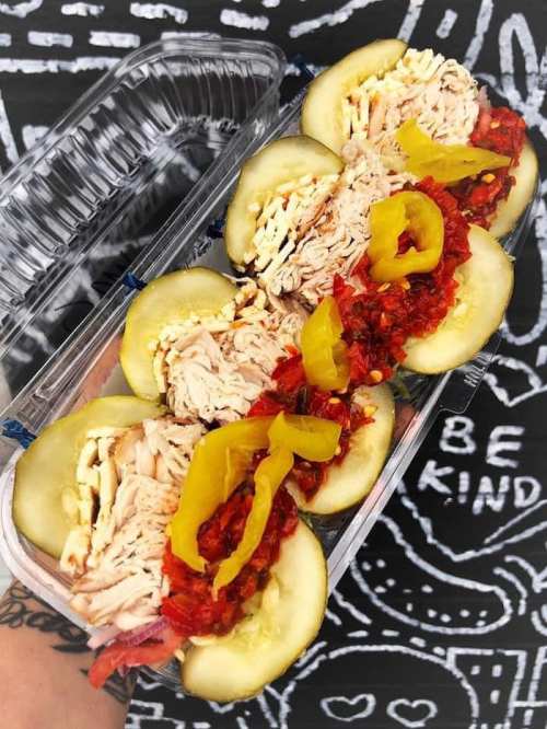 A hand holding a container with sliced pickles filled with shredded chicken, peppers, and a red sauce.