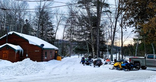 A snowy landscape featuring a wooden cabin, snowmobiles parked nearby, and trees in the background at dusk.