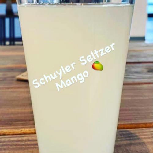 A glass of mango seltzer on a wooden table, with the text "Schuyler Seltzer Mango" overlaying the image.