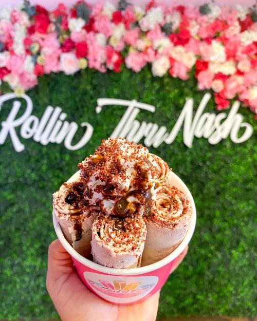 A hand holding a cup of rolled ice cream topped with chocolate sauce and sprinkles, with a floral backdrop.