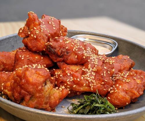 A plate of crispy chicken wings coated in red sauce and sesame seeds, served with a dipping sauce and garnished with greens.