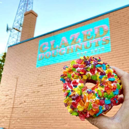 A colorful donut topped with cereal held in front of a wall featuring "Glazed Doughnuts" sign.