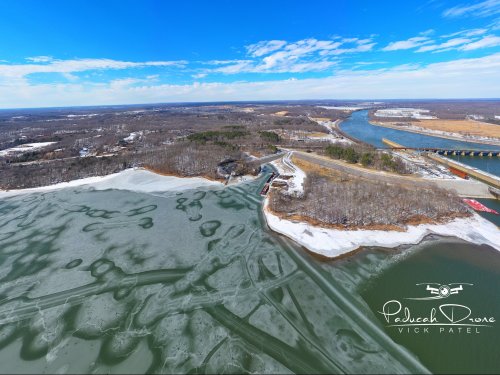 Aerial view of a river with ice and snow, surrounded by trees and a road under a clear blue sky.