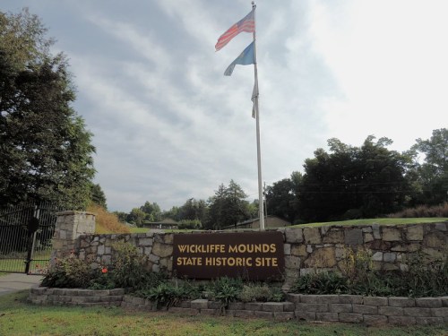 Sign for Wickliffe Mounds State Historic Site, featuring flags and surrounded by greenery under a cloudy sky.