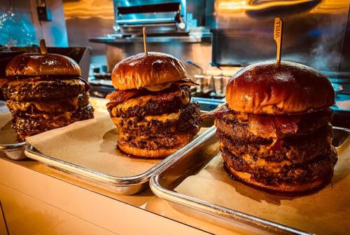 Three stacked burgers with bacon and melted cheese on toasted buns, served on metal trays in a restaurant kitchen.