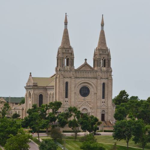 A large stone church with two tall spires, surrounded by trees and a grassy area under a cloudy sky.