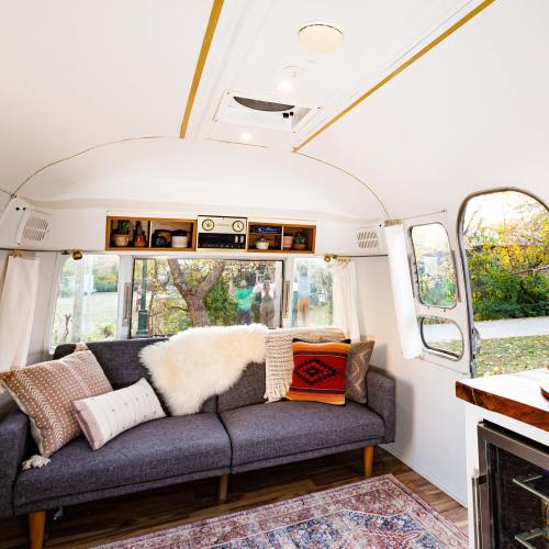 Cozy interior of a vintage trailer with a sofa, decorative pillows, and large windows showcasing a natural view outside.