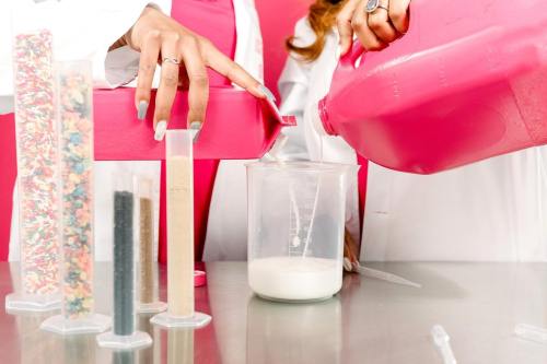 A person in a lab coat pours pink liquid from a jug into a measuring cup, surrounded by colorful ingredients in test tubes.
