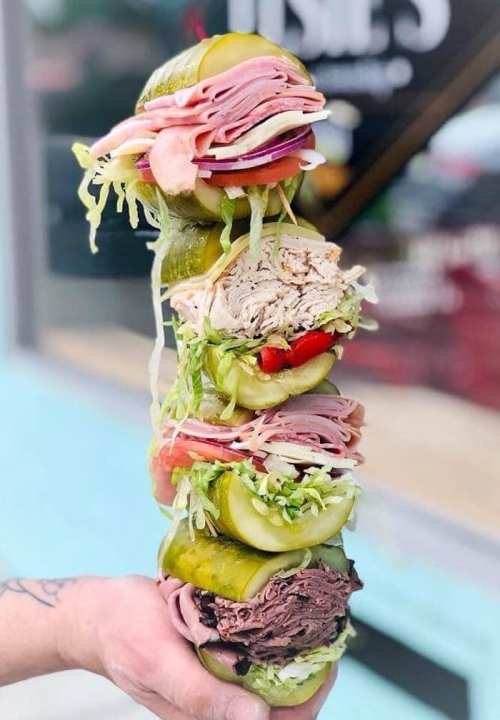 A towering sandwich stacked with layers of meats, pickles, lettuce, and tomatoes, held by a hand.