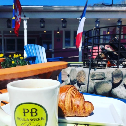 A cup of coffee and a croissant on a table, with a fire pit and flags in the background on a sunny day.