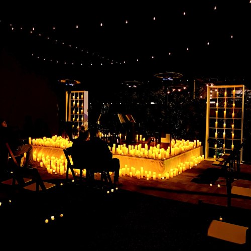 A cozy outdoor space illuminated by numerous candles, with people seated and string lights overhead.