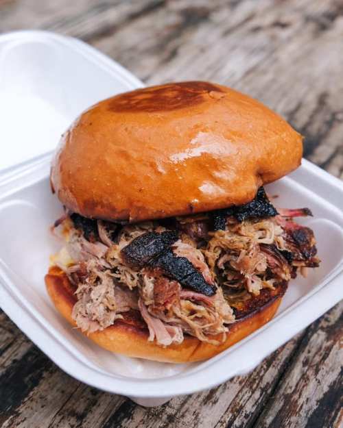 A close-up of a pulled pork sandwich on a soft bun, served in a takeout container.