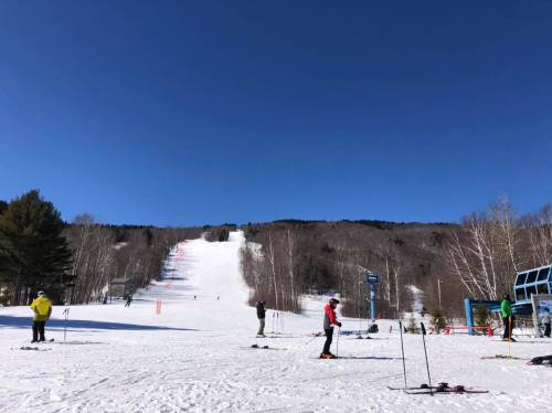Skiers on a snowy slope under a clear blue sky, with trees and a ski lift in the background.