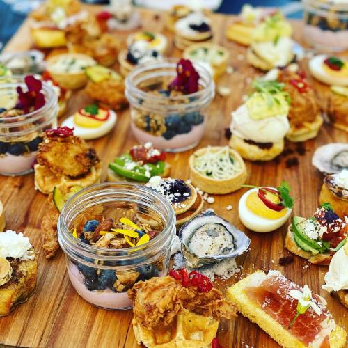 A wooden platter filled with various gourmet appetizers, including jars of food, oysters, and colorful bite-sized treats.