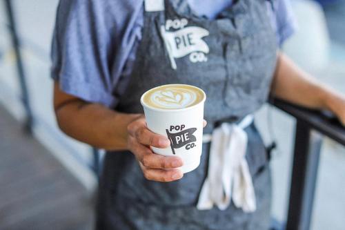A person in an apron holds a coffee cup with a latte art design, featuring the logo "Pop Pie Co." on the cup.