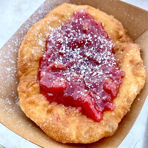 A golden fried pastry topped with red fruit jam and dusted with powdered sugar, served in a paper tray.