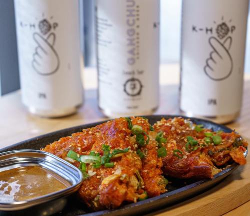 A sizzling plate of spicy chicken wings topped with green onions, accompanied by a dipping sauce and drinks in the background.