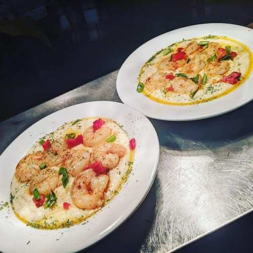 Two plates of shrimp and grits, garnished with green onions and red peppers, served on a silver surface.