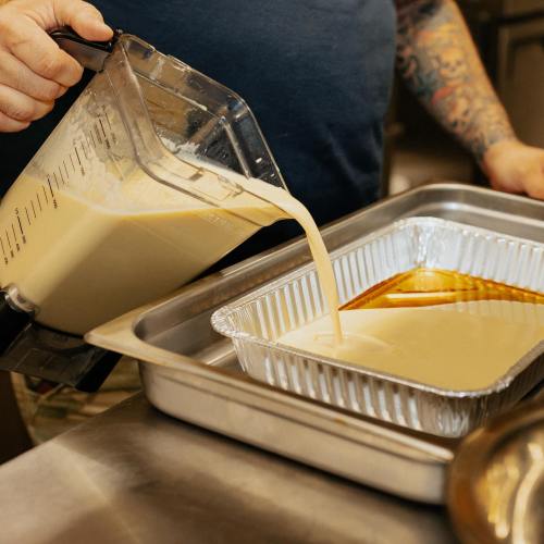 A person pours a creamy mixture from a blender into a metal tray in a kitchen setting.