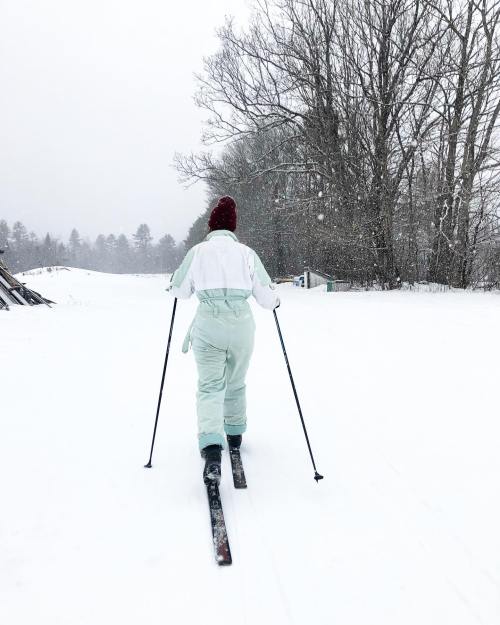 A person skiing on a snowy trail, wearing a light green ski suit and a red hat, surrounded by trees in winter.