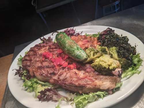 A grilled steak served on a bed of greens, accompanied by broccoli, beans, and a jalapeño pepper.