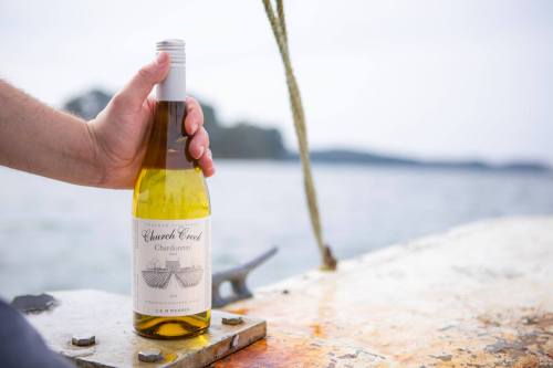 A hand holding a bottle of Church Creek Chardonnay on a boat, with a scenic water view in the background.