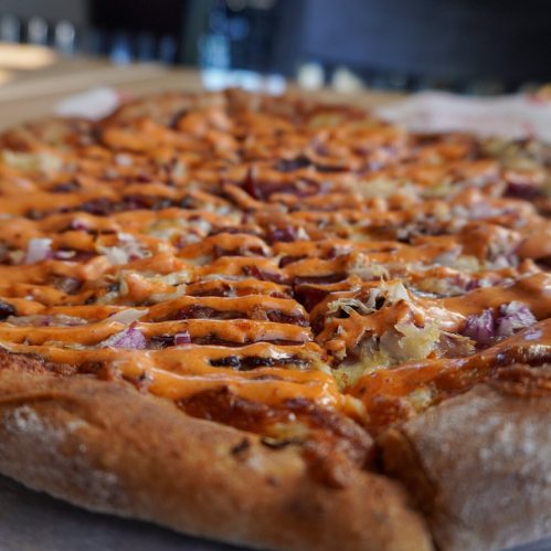 Close-up of a pizza topped with drizzled sauce, red onions, and a golden crust, set on a wooden surface.