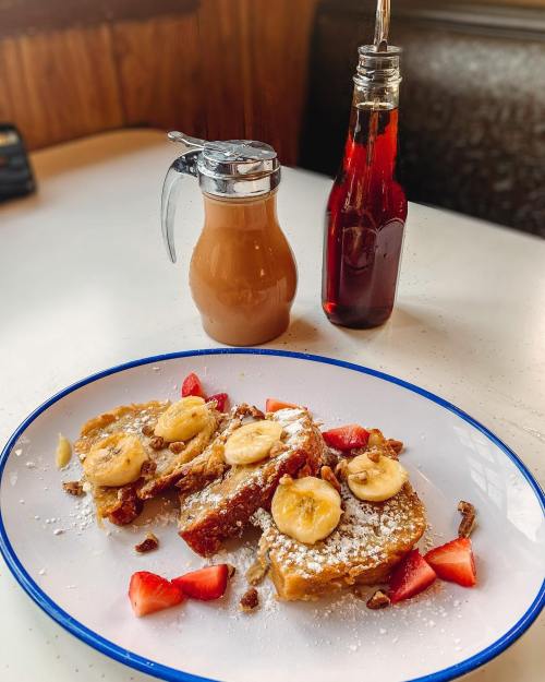 A plate of French toast topped with banana slices and strawberries, with syrup and a drink on the side.