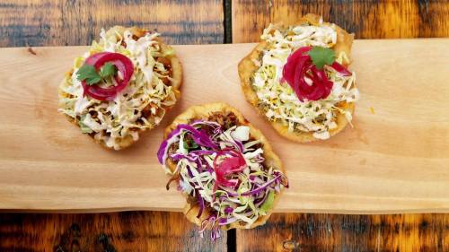 Three colorful tostadas topped with shredded cabbage, pickled onions, and cilantro on a wooden platter.