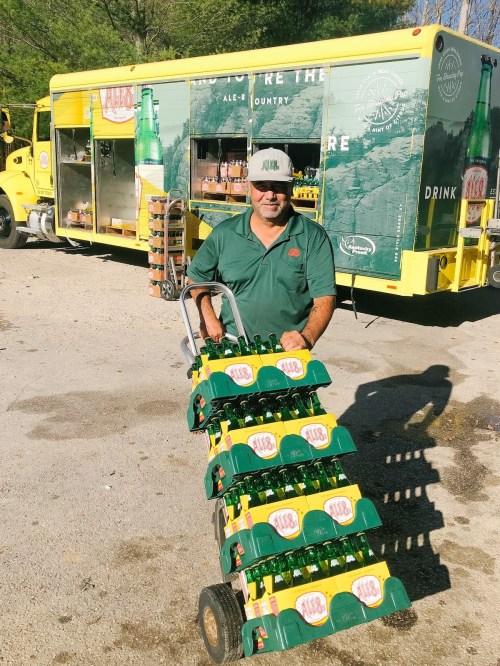 A man in a green shirt and cap pushes a cart loaded with bottles in front of a colorful beverage truck.