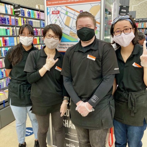 Four employees in black shirts and masks pose together in a bookstore, smiling and showing peace signs.