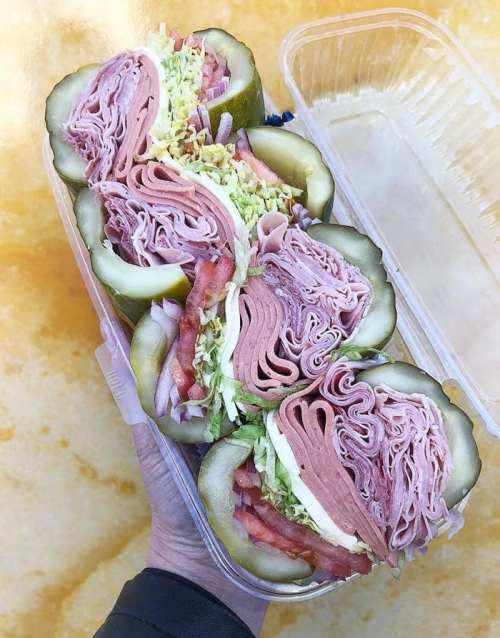 A hand holding a clear container filled with pickle halves stuffed with layers of deli meats, lettuce, and tomatoes.