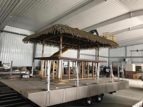 A wooden structure with a thatched roof, set on a trailer inside a large warehouse.