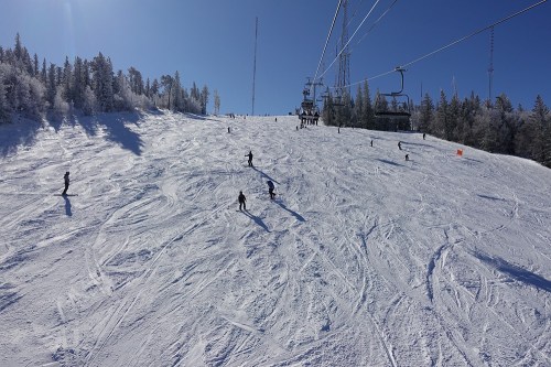 Skiers navigate a snowy slope under a clear blue sky, with a ski lift in the background and trees lining the hill.