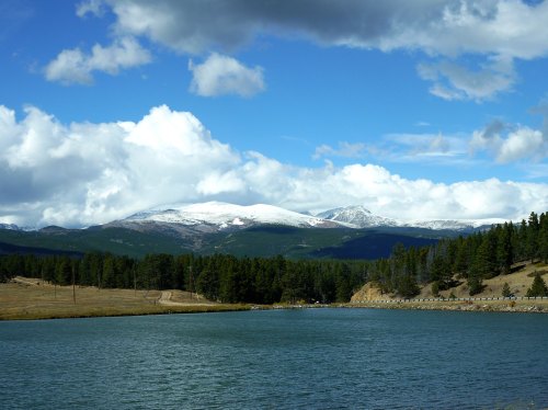 Scenic view of a lake with mountains in the background, partially covered in snow, under a blue sky with clouds.