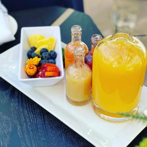 A platter with a large jug of orange juice, small bottles of various juices, and fresh fruit including pineapple and berries.