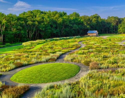 Aerial view of a landscaped garden with winding paths, circular grass areas, and a rustic barn surrounded by trees.